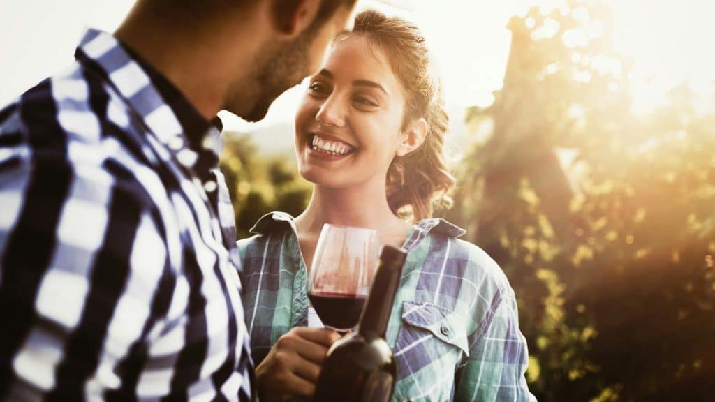 A smiling woman holding a glass and bottle of wine while looking at a man outdoors.
