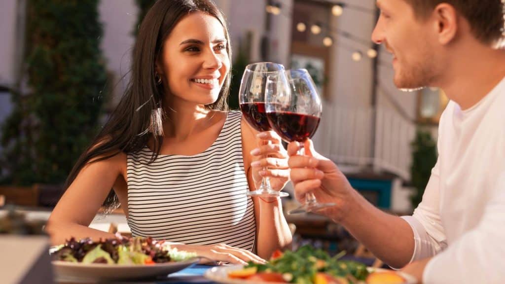 A couple smiling and clinking glasses of red wine during a meal.