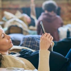 A woman using her smartphone while lying on a couch with a man in the background.