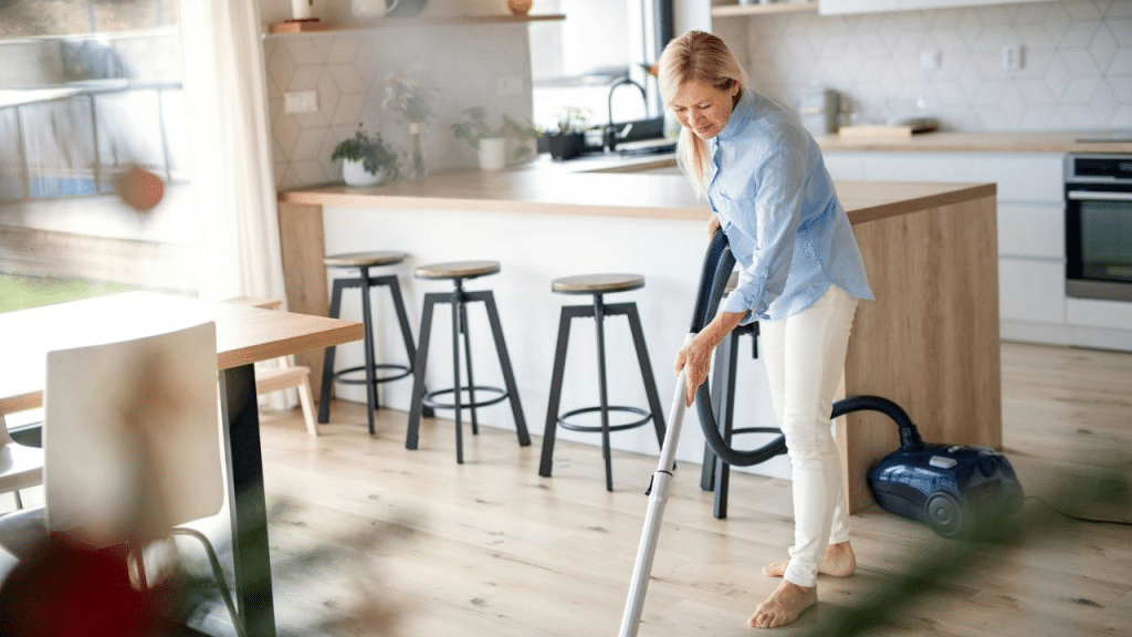 A smiling woman vacuums a bright, modern kitchen and dining room while barefoot.