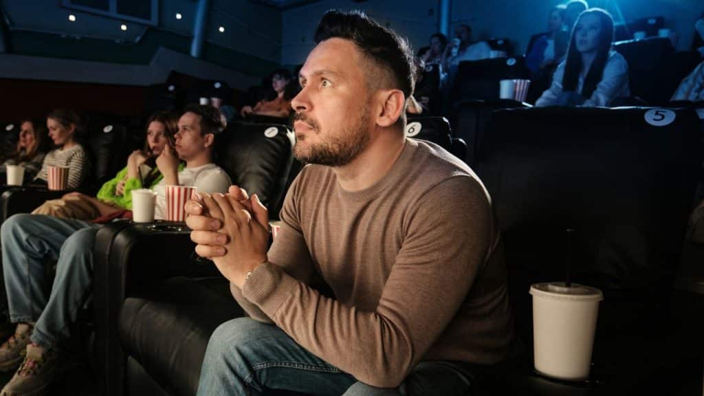 A man with his hands clasped sits and watches a movie in a dark theater.