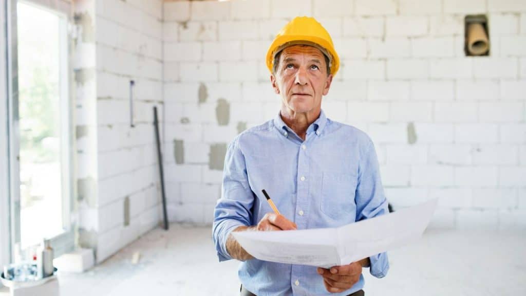 A senior man in a hard hat looks up, holding a blueprint and a pencil.