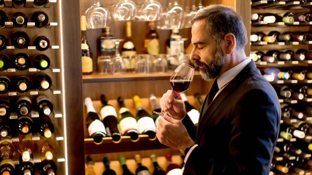 A man in a suit sniffs a glass of red wine in a wine cellar.