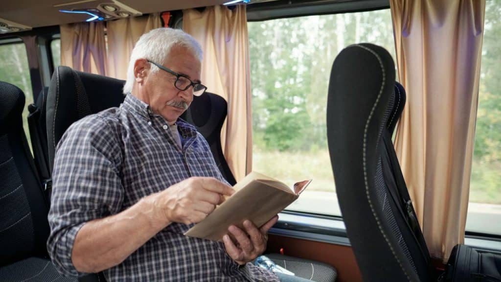 A mature man in glasses and a flannel shirt reads a book on a bus.