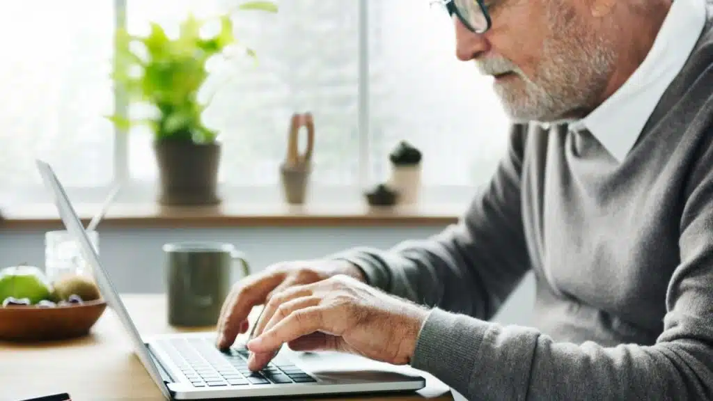 A side view of a senior man typing on a laptop with a mug nearby.