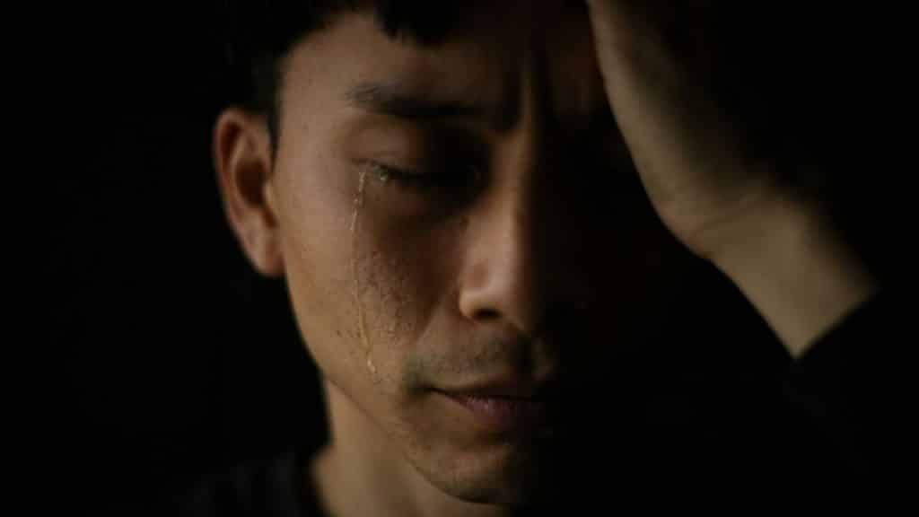 A crying man in the dark holds his hand to his head.