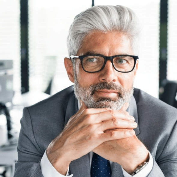 A senior businessman with gray hair and a beard is wearing glasses and sitting at a desk.
