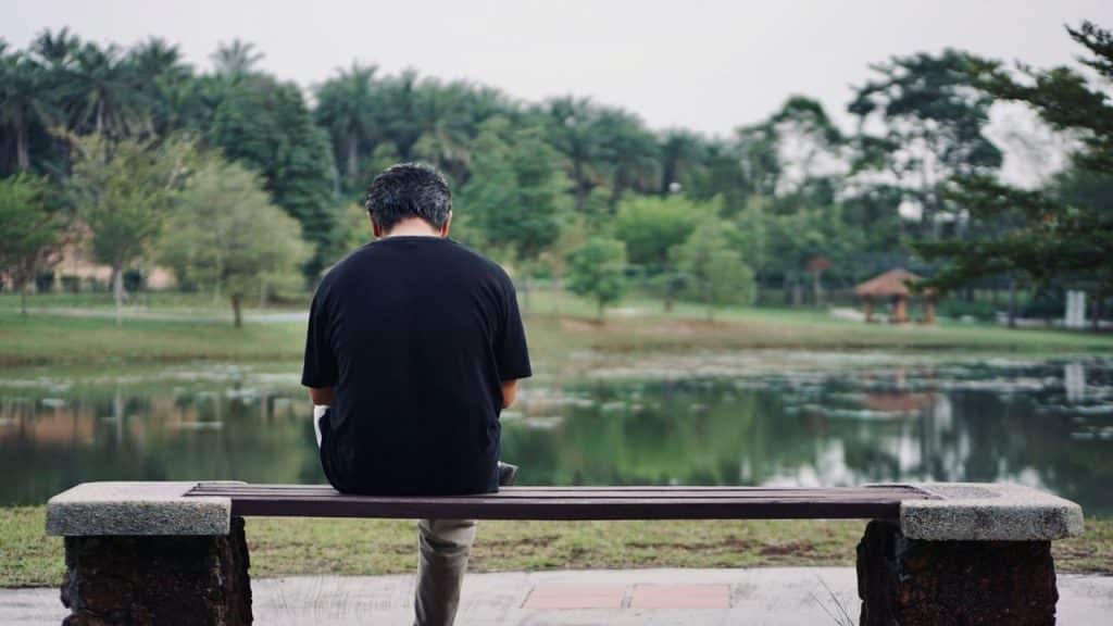 A person with their back to the camera sits on a bench, looking at a lake.