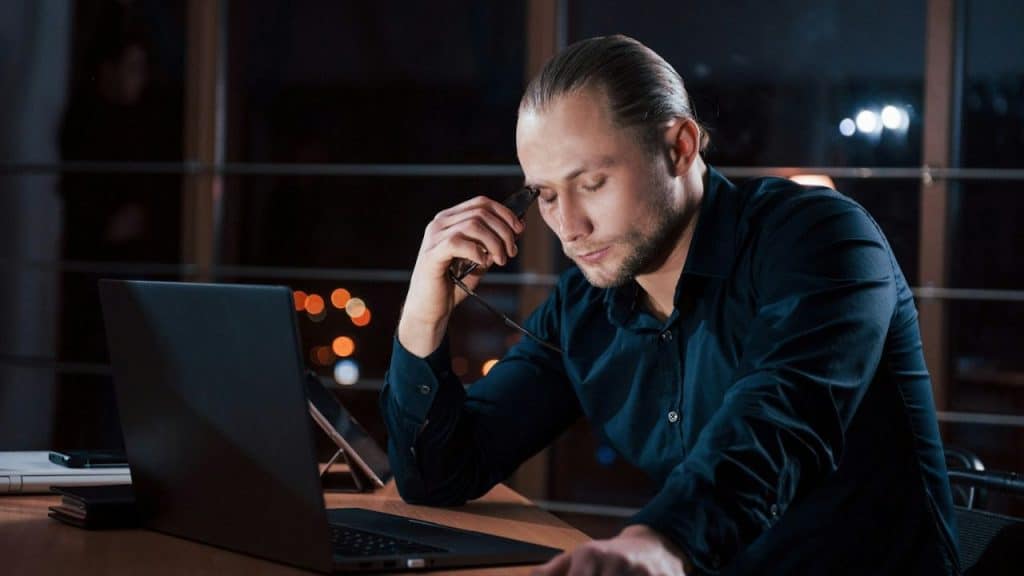 A man sits at a desk in the dark, eyes closed, holding his glasses.