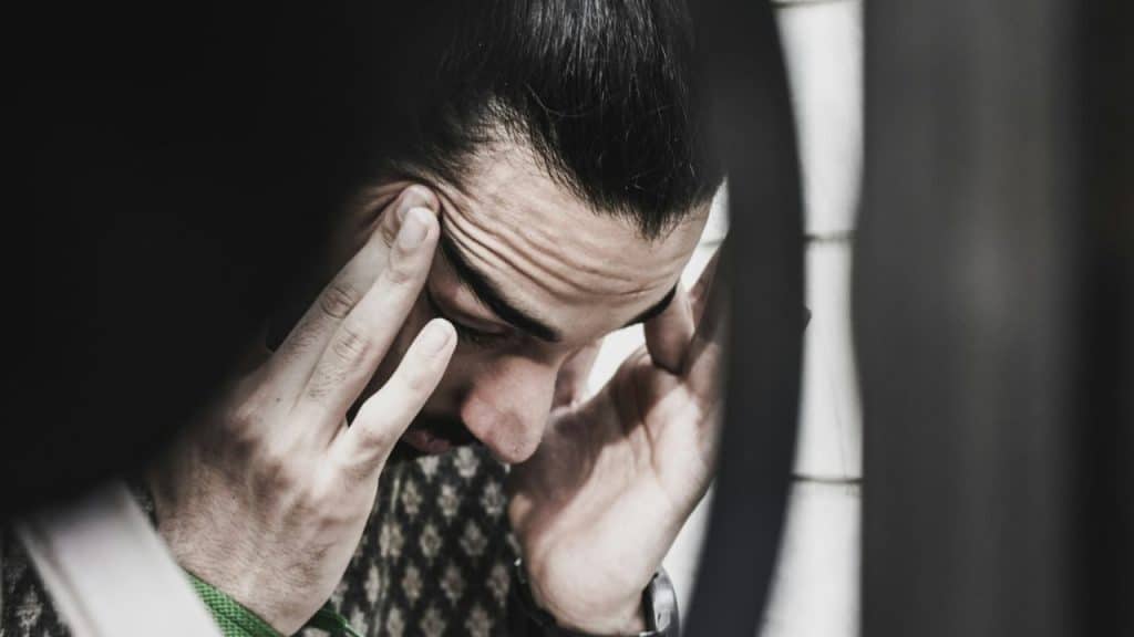 A man with a pained expression and his eyes closed, touches his temples.