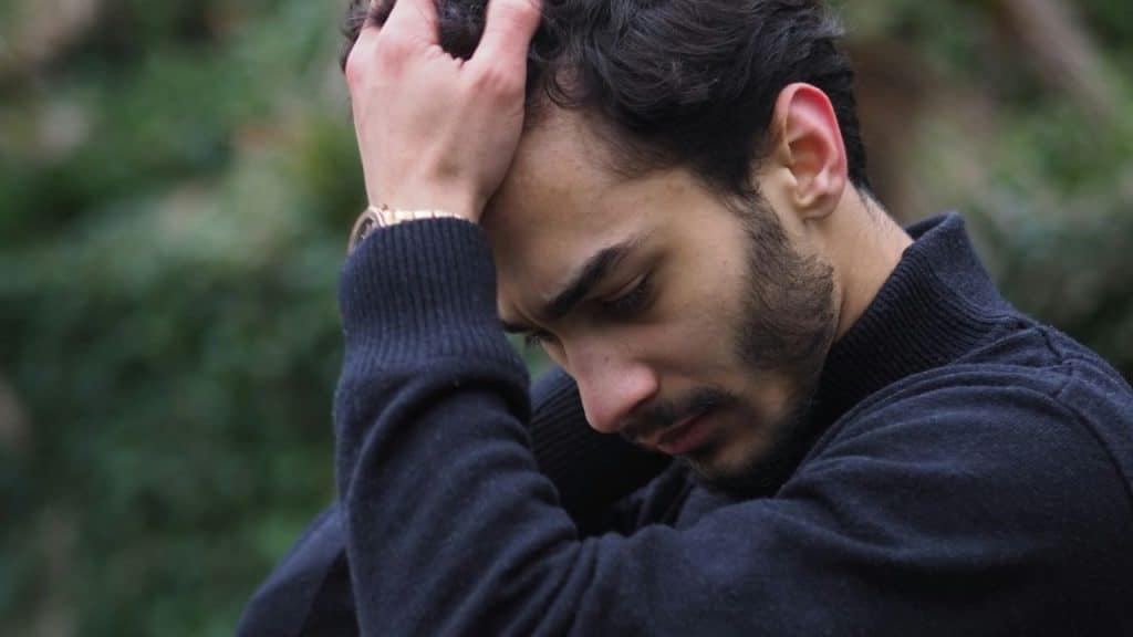 A man with dark hair and a beard sits outdoors with his hands on his head, looking down.