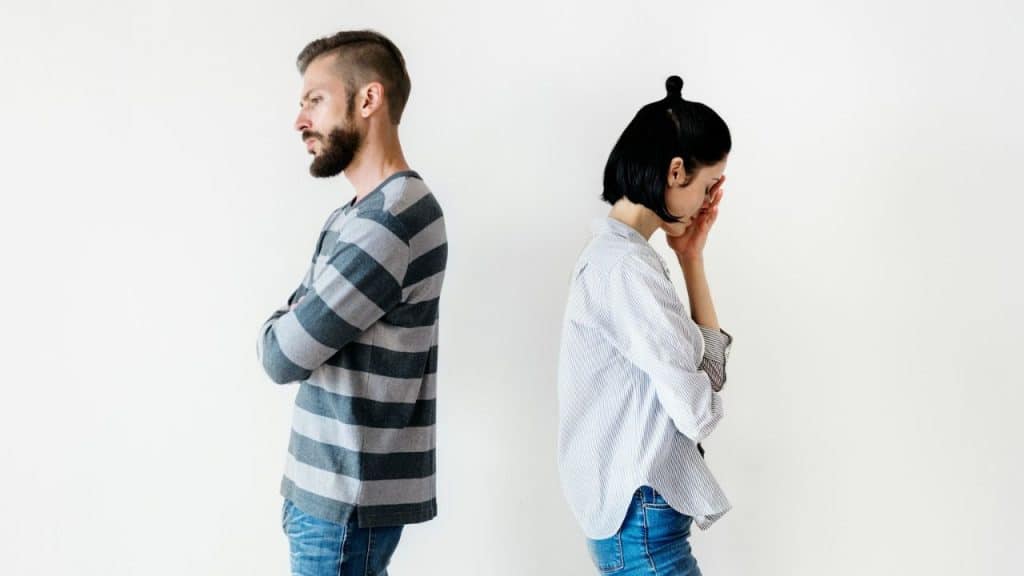 A man and woman stand back to back against a white wall.