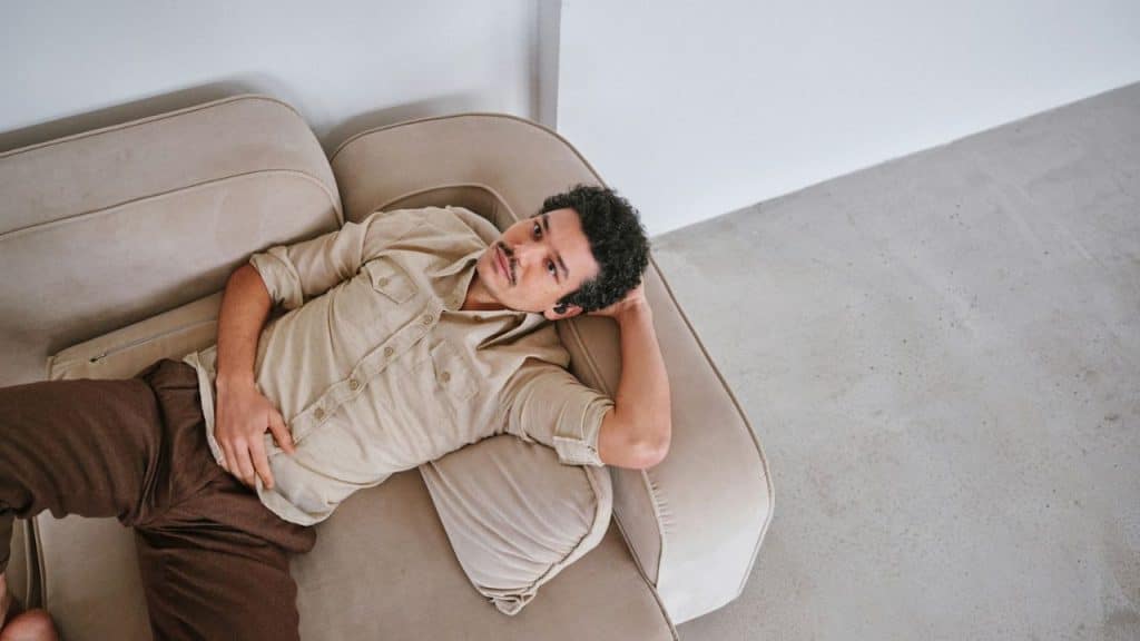 A man with dark, curly hair and a mustache is lying on a light-colored couch.
