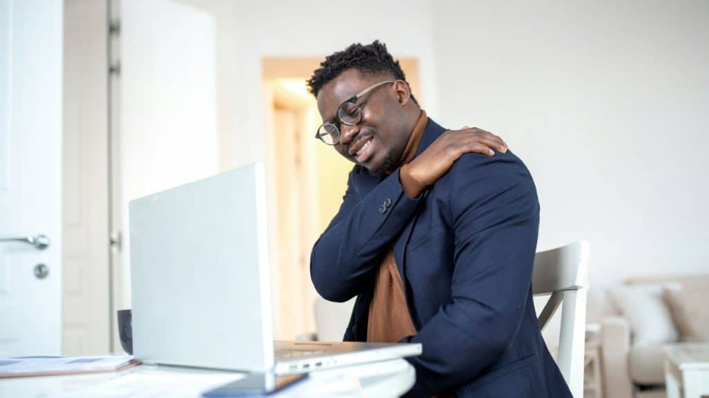 A Black man in glasses and a dark suit jacket sits at a table, massaging his shoulder.