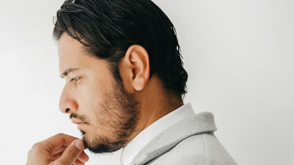 A man with a beard and dark hair looks down and to the side, touching his chin.