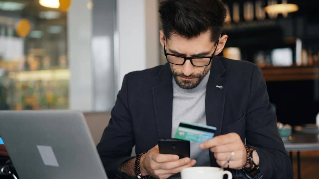 A man with a beard and glasses is sitting at a cafe table, holding a phone and a credit card. He appears to be making an online transaction.
