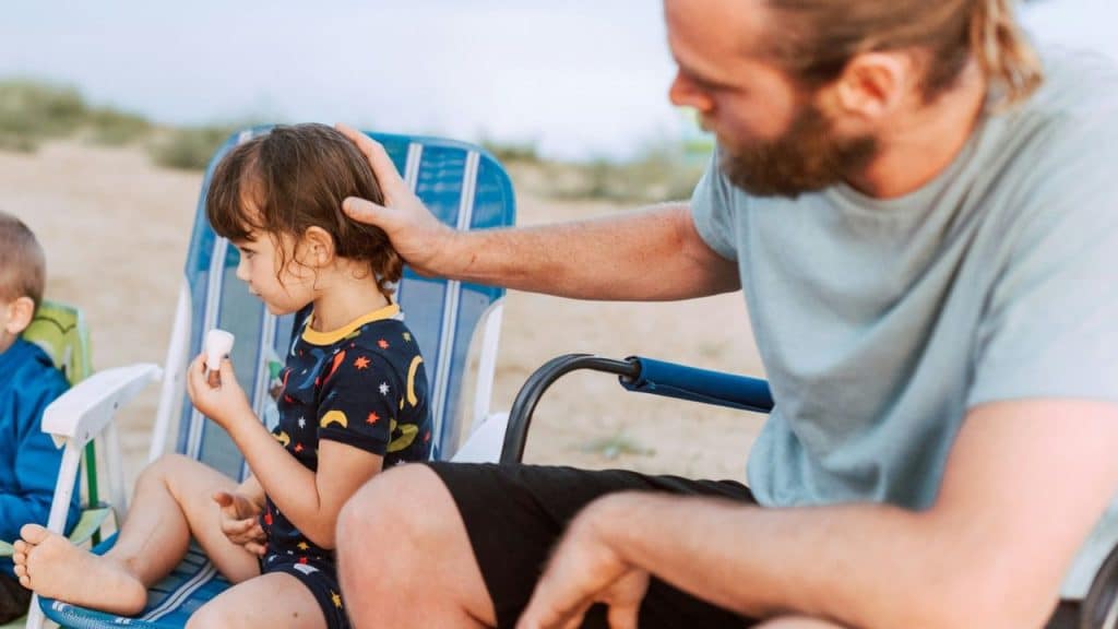 A man with a beard sits next to a child in a beach chair, holding the back of the child's head.