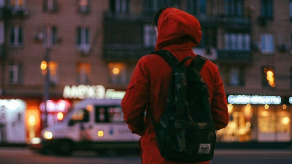 A person wearing a red hooded jacket and a camouflage backpack is standing on a street at dusk, with their back to the camera.