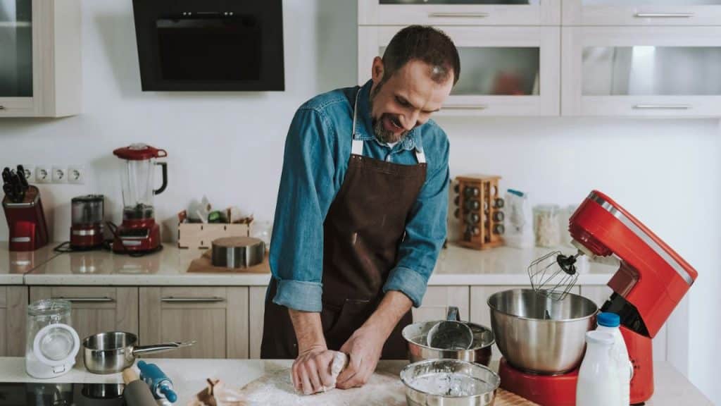 A man with a beard and an apron is kneading dough on a countertop in a kitchen.