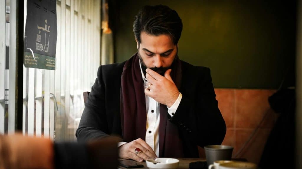 A man with a beard and a black jacket is sitting at a cafe table, with a worried look on his face, holding his chin.