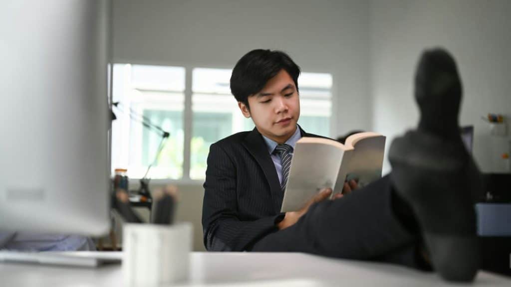 A man in a suit reads a book with his feet up on a desk.