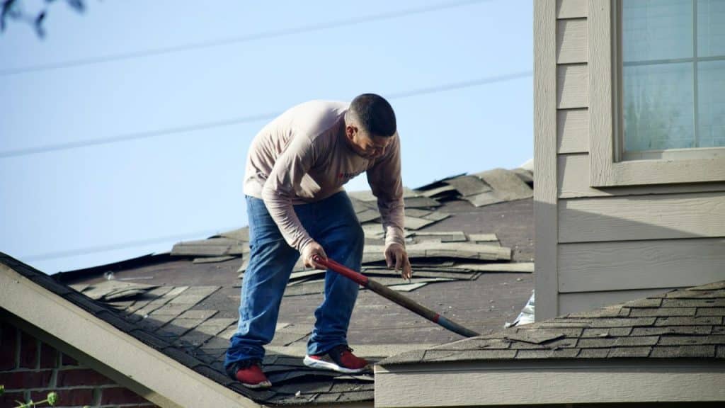 A man on a roof uses a shovel to work on damaged shingles.