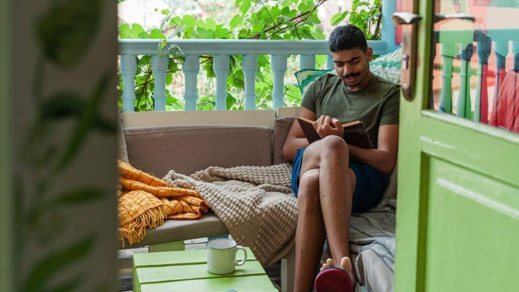 A smiling man with a mustache sits on an outdoor sofa, reading a book.