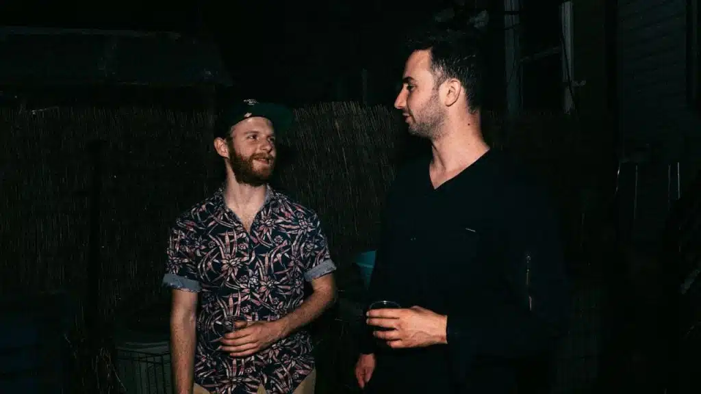 Two men stand outside at night, talking. One man wears a patterned shirt and a backwards hat, and the other wears a black shirt.