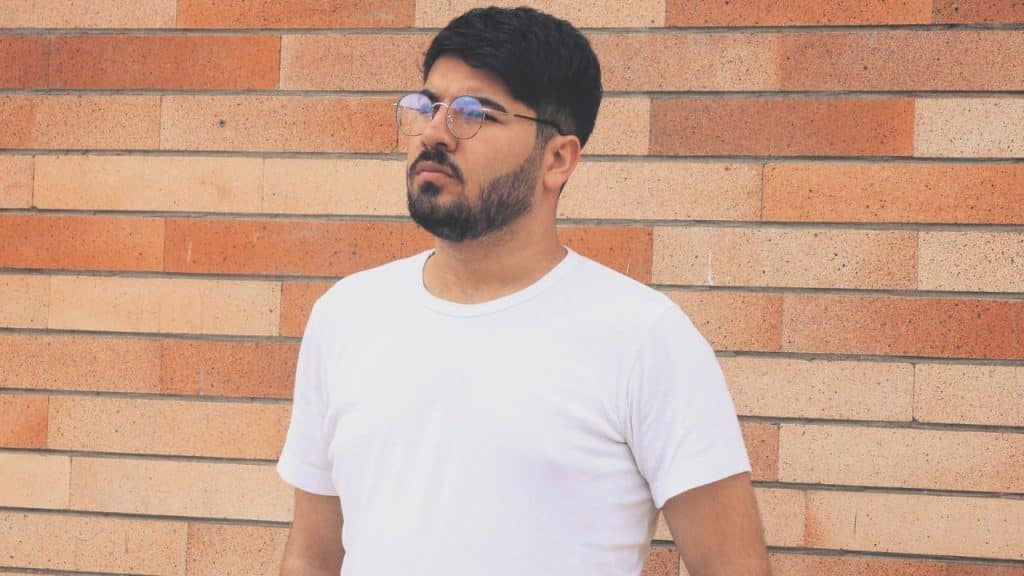 A bearded man wearing a white t-shirt and glasses stands in front of a tan brick wall.