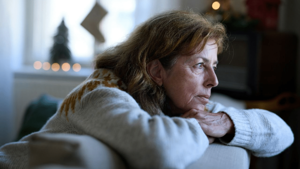 A mature woman in a white sweater looks off into the distance, with her head resting on her hands.