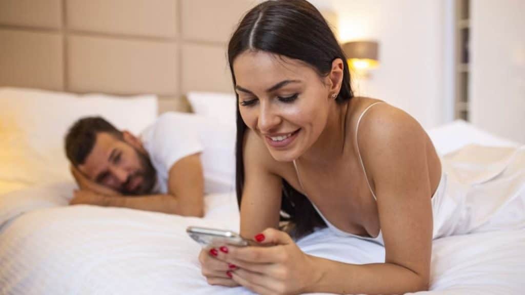 A wife in bed scrolling on her phone while her husband is just looking at her