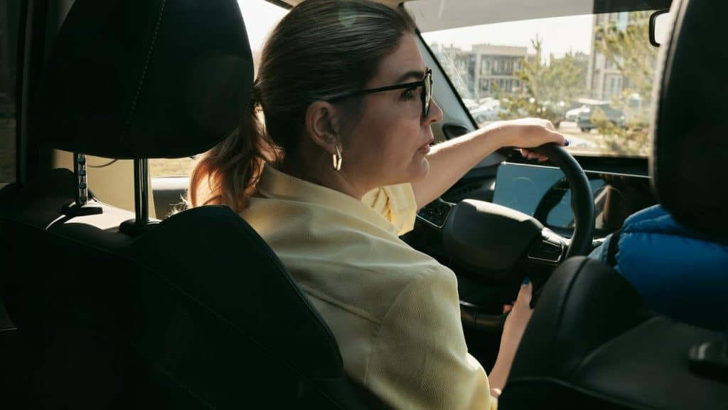A woman in glasses drives a car while looking back over her shoulder.