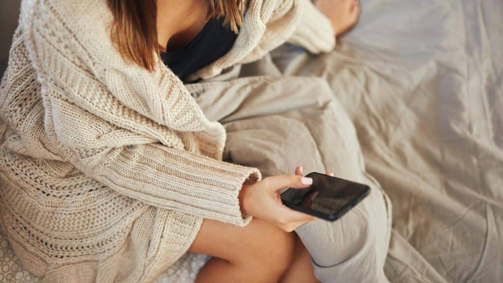 A woman in a cozy sweater sits on a bed holding a smartphone.