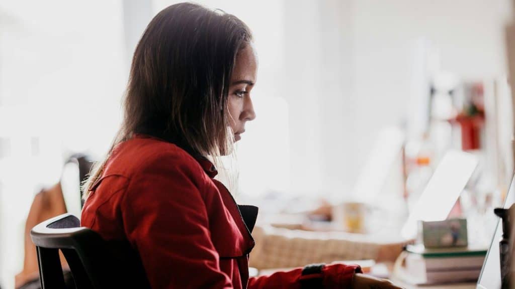 A woman in a red jacket works at a desk while focused on her laptop.