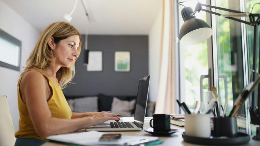 A woman in a yellow top works on a laptop at a desk by the window.