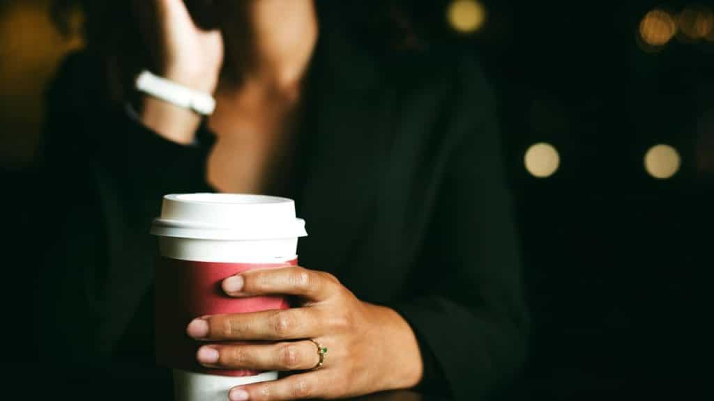 A person in a dark outfit holds a takeaway coffee cup with a red sleeve.