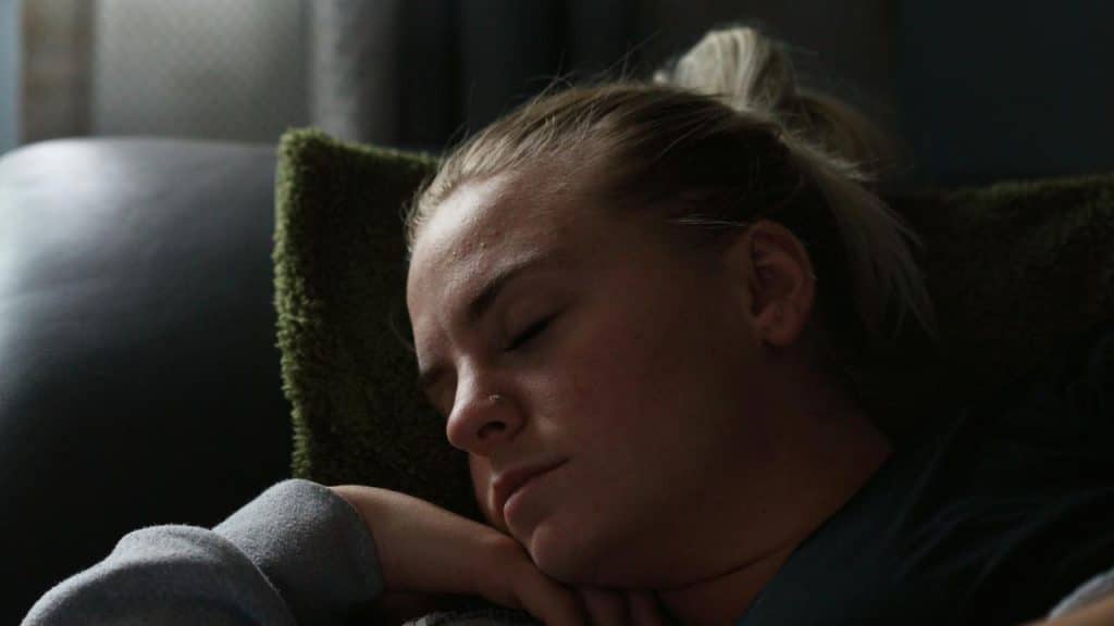 A woman rests on a couch with her eyes closed, lying against a pillow.