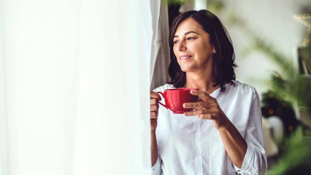 A woman in a white shirt holds a red mug while looking out the window with a smile.