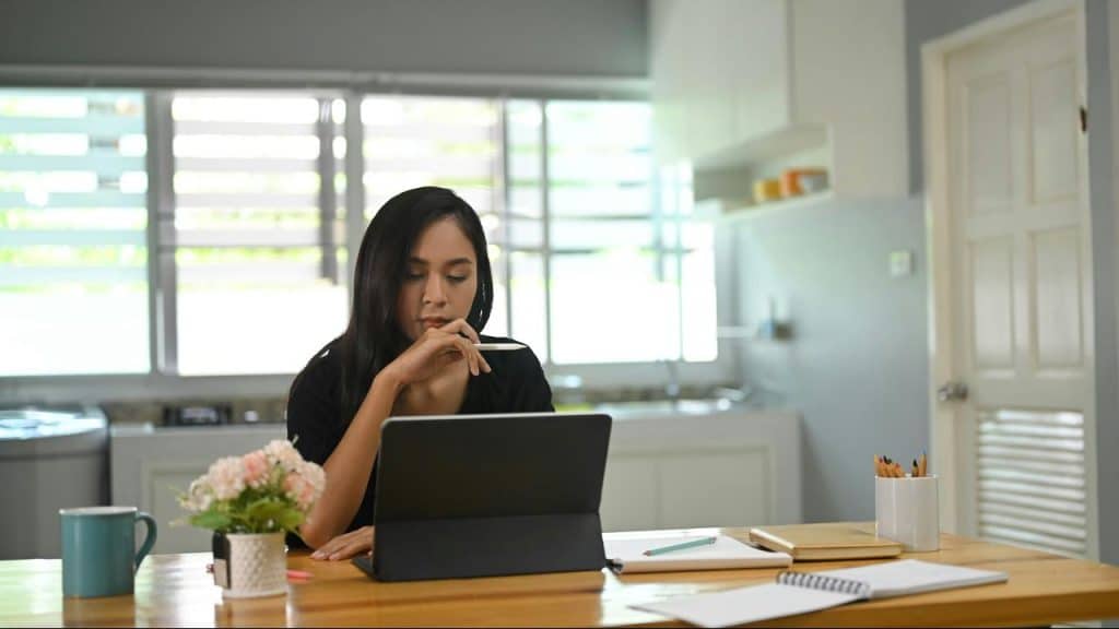 A woman sits at a table in her kitchen working on a tablet with notes and coffee nearby.