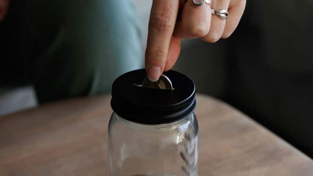 A person putting a coin into a glass jar with a slot lid.