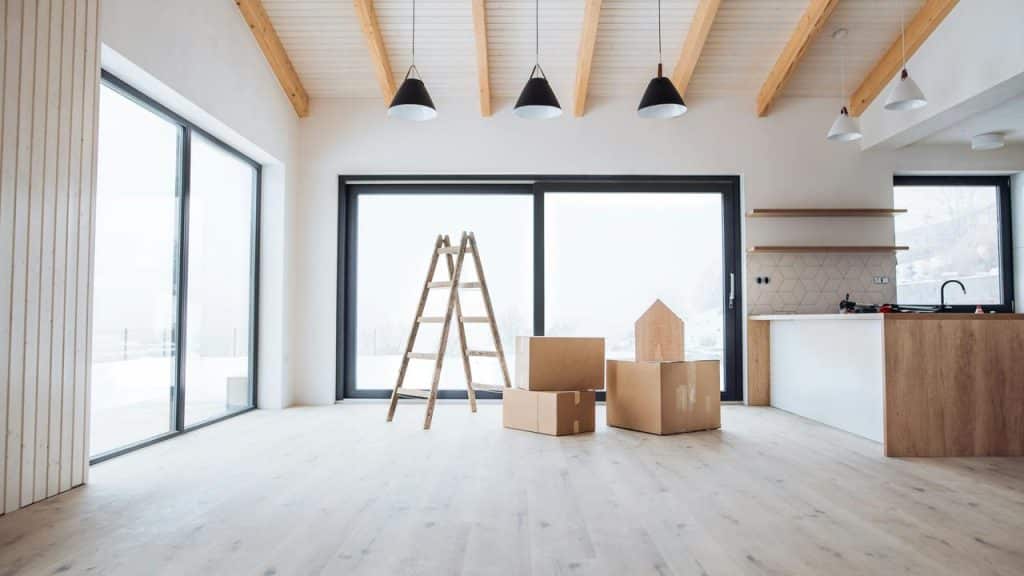 A modern empty room with moving boxes and a wooden ladder near large windows.