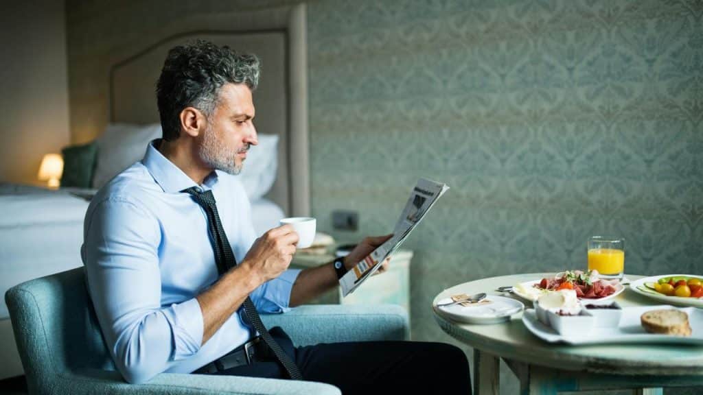 A man in a shirt and tie reading a newspaper while drinking coffee at a breakfast table.
