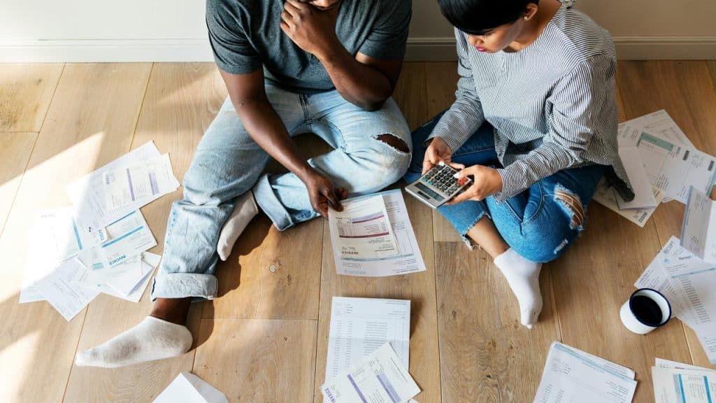 A couple sitting on the floor surrounded by bills and invoices while using a calculator.