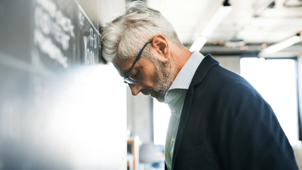 A middle-aged man with gray hair, a beard, and glasses leans his head against a black wall.