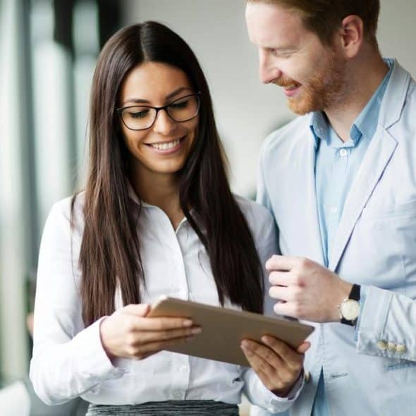 A woman in glasses and a man in a blazer smiling while looking at a tablet.