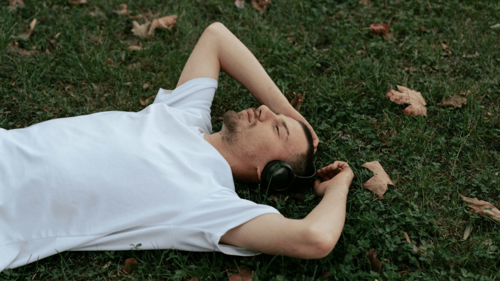 A man in a white T-shirt lies in the grass with headphones on.