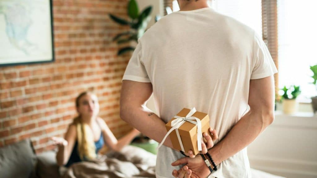 A man hides a gift box behind his back as a woman sits on a bed looking upset.