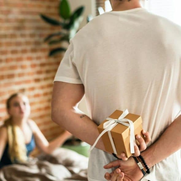 A man hides a gift box behind his back as a woman sits on a bed looking upset.