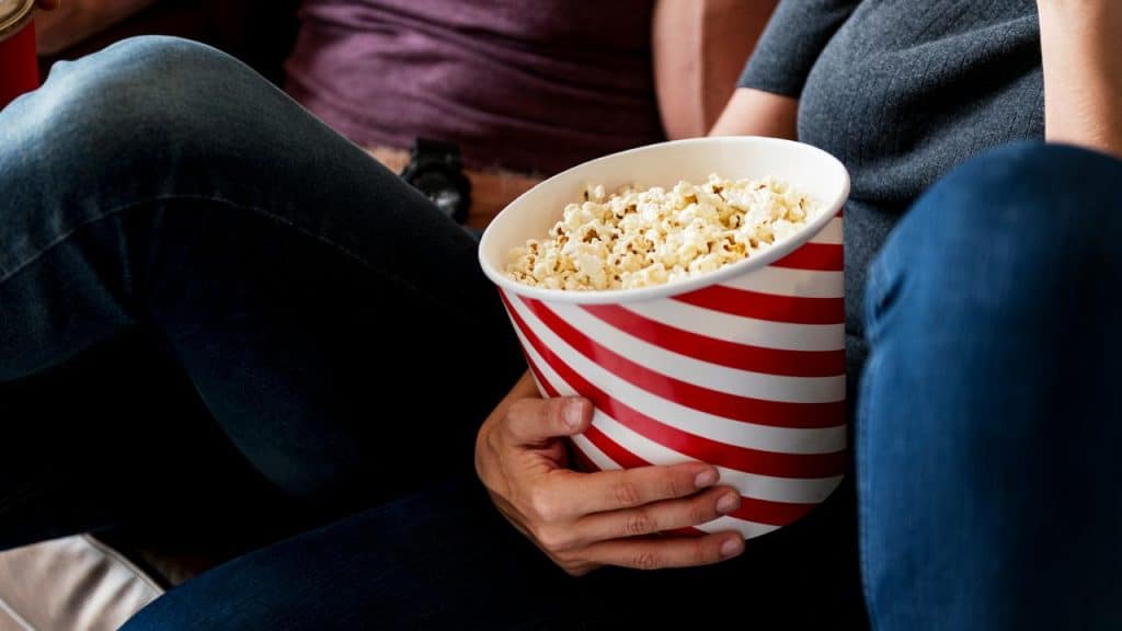 A person holds a large striped bucket of popcorn while sitting close to someone.