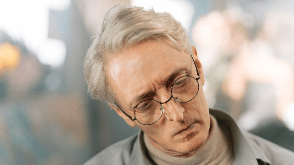 A middle-aged man with gray hair and glasses looks downward with a sad expression.